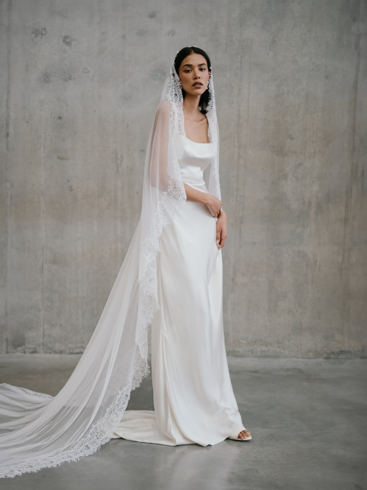 Side-angle standing portrait of the Clara wedding gown and long draped lace veil at Carte Blanche Bride.